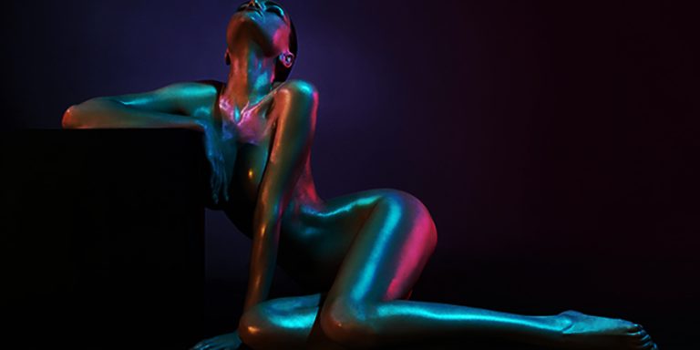 Mujer desnuda iluminada con luces neón azul y rosa en pose sensual sobre fondo oscuro