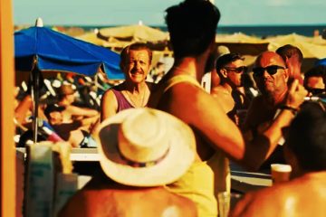 Hombre discutiendo en un chiringuito de playa abarrotado en Maspalomas, con bañistas y sombrillas al fondo bajo el sol.