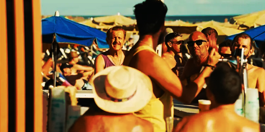 Hombre discutiendo en un chiringuito de playa abarrotado en Maspalomas, con bañistas y sombrillas al fondo bajo el sol.