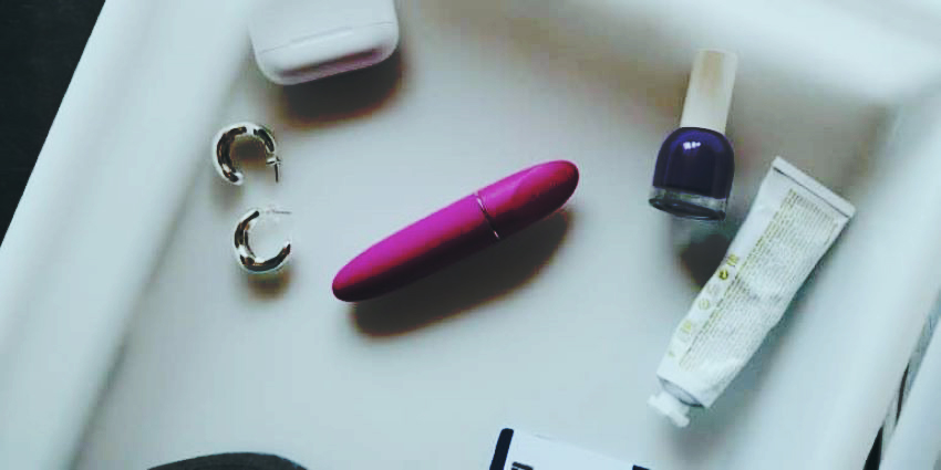 Vibrador discreto rosa de tamaño compacto junto a objetos personales sobre bandeja blanca.