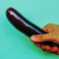 Mano sujetando una berenjena sobre fondo turquesa como metáfora visual del tamaño del pene