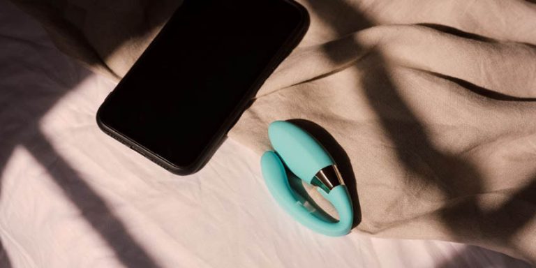 Vibrador para parejas y smartphone sobre una cama, representando la intimidad en las relaciones a distancia mediante tecnología interactiva.